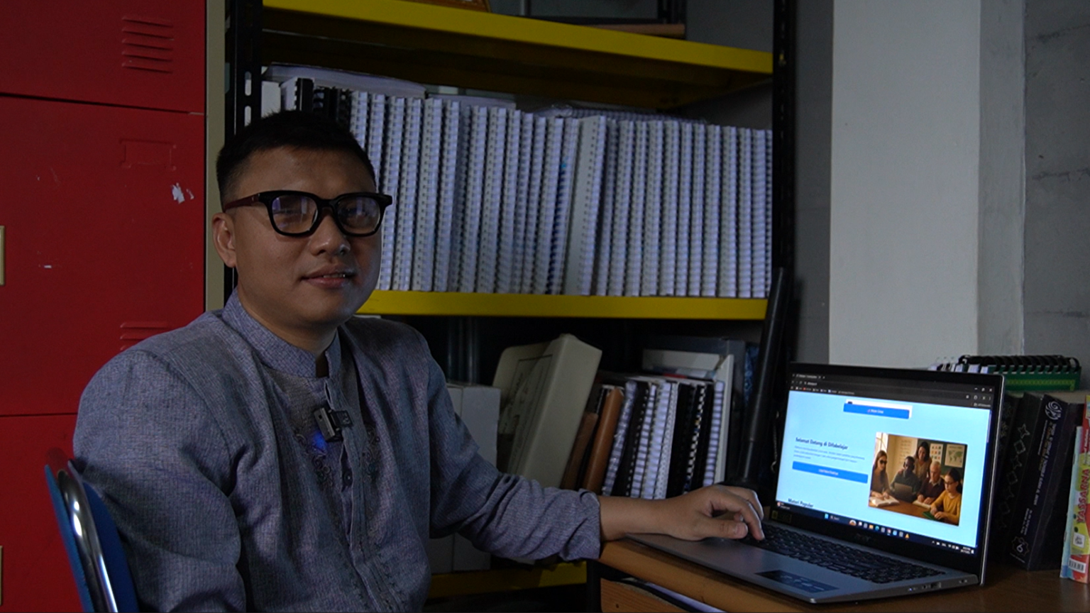 Sou um homem adulto, de pele morena clara, com cabelo preto curto e uso óculos de armação escura. Estou sentado em uma cadeira, em um ambiente que parece um escritório ou sala de estudos. Visto uma camisa de manga longa em tom azul acinzentado. À minha frente, sobre uma mesa, há um notebook aberto, e minha mão direita está apoiada próximo ao teclado. Na tela do computador aparece um site com fundo claro e uma imagem de pessoas reunidas. Ao fundo, há estantes metálicas com várias pastas e cadernos organizados, além de um armário vermelho à esquerda. O ambiente é bem iluminado e organizado.