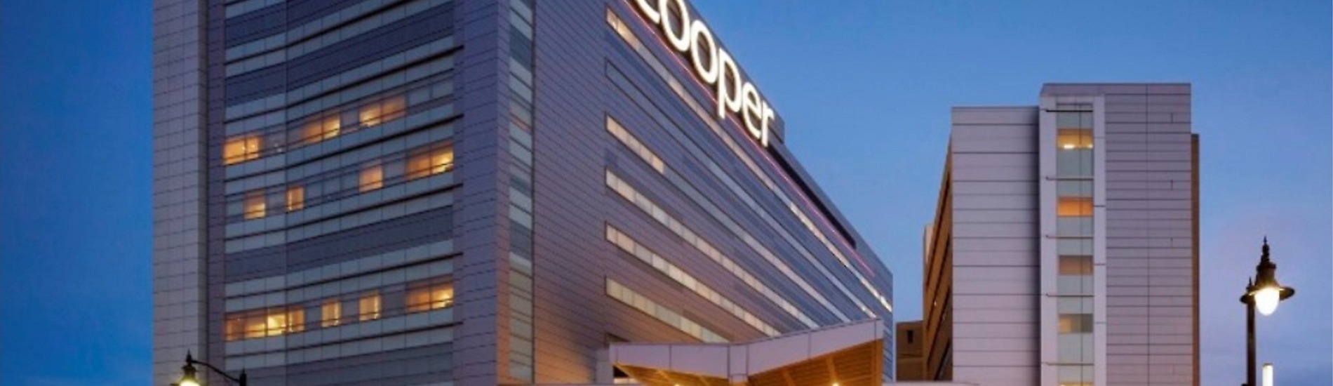 Imagem externa de um grande edifício hospitalar moderno, visto ao entardecer. A construção é alta, com fachada em tons de cinza e linhas retas, composta por vários andares com janelas horizontais iluminadas por luz amarela. No topo do prédio, há um letreiro luminoso com a palavra “Cooper”, parcialmente visível. À frente, nota-se uma estrutura de entrada com cobertura. O céu ao fundo está azul-escuro, indicando início da noite, e há postes de iluminação pública acesos nas laterais da imagem.