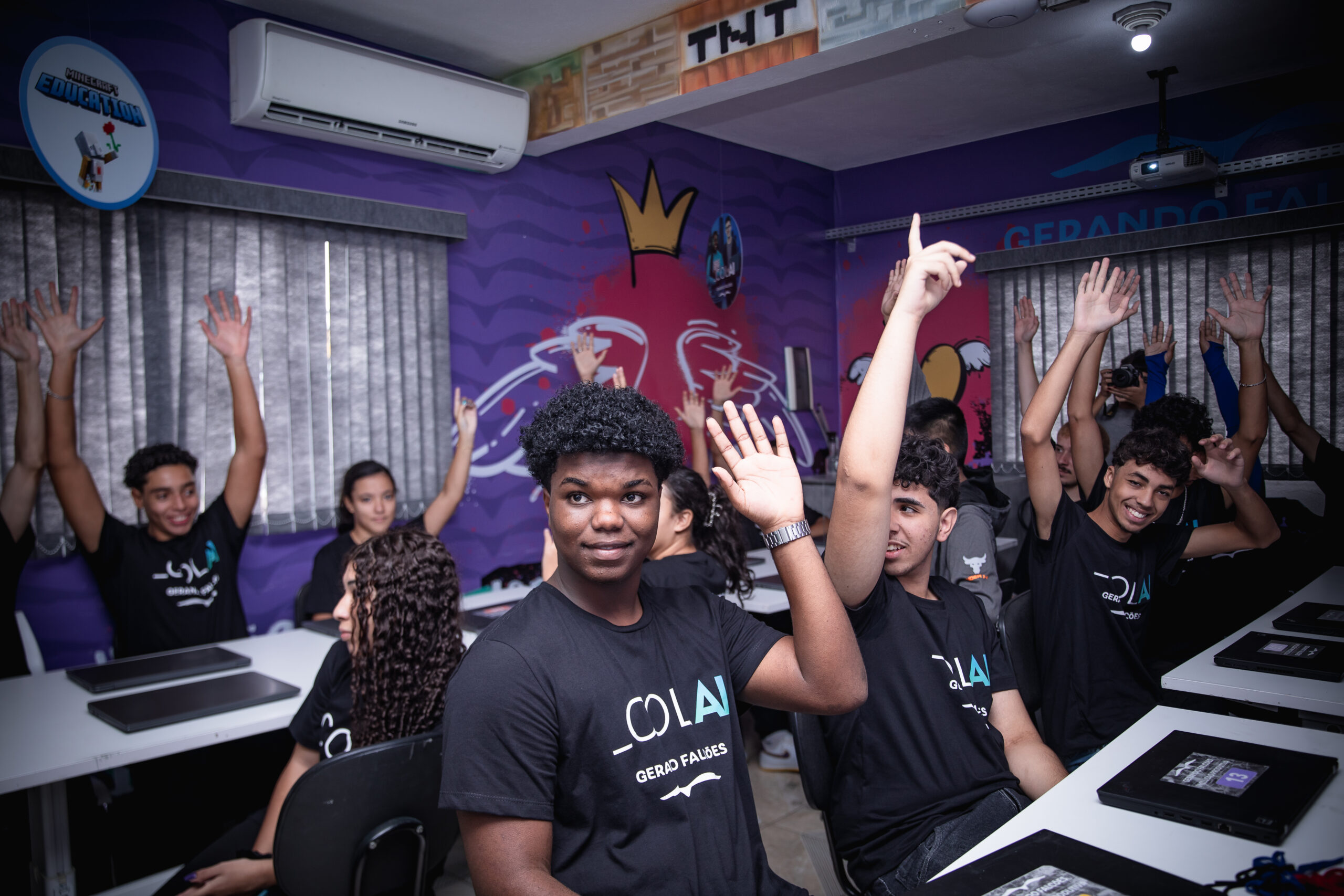 Sala de aula com paredes roxas decoradas com grafites e elementos visuais coloridos. No ambiente, cerca de dez adolescentes estão sentados em mesas com notebooks fechados à frente. A maioria veste camisetas pretas com a palavra “COLAI” estampada. Vários estudantes levantam a mão, demonstrando participação e entusiasmo. Em primeiro plano, um jovem de cabelo curto e crespo também levanta a mão e olha para frente com expressão atenta. Ao fundo, há cortinas cinzas, um projetor preso ao teto e um aparelho de ar-condicionado na parede. O ambiente transmite clima de atividade educacional dinâmica e colaborativa.