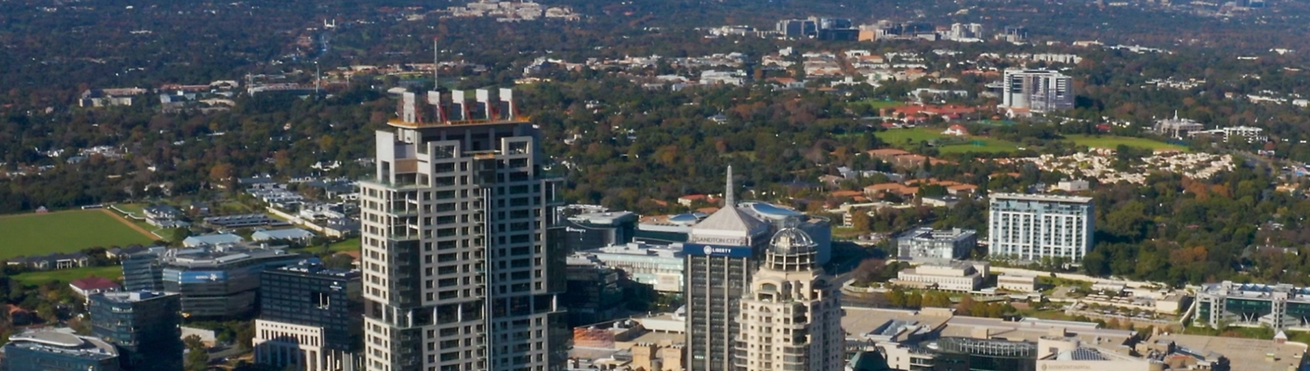 Imagem aérea de uma área urbana durante o dia. Em primeiro plano, há edifícios altos e modernos, com fachadas claras e telhados planos. Ao redor, outros prédios comerciais e residenciais de diferentes alturas. No fundo, a paisagem se estende com muitas áreas verdes, árvores e bairros de casas baixas, formando um contraste entre a região mais verticalizada e áreas arborizadas. O céu não aparece na imagem.