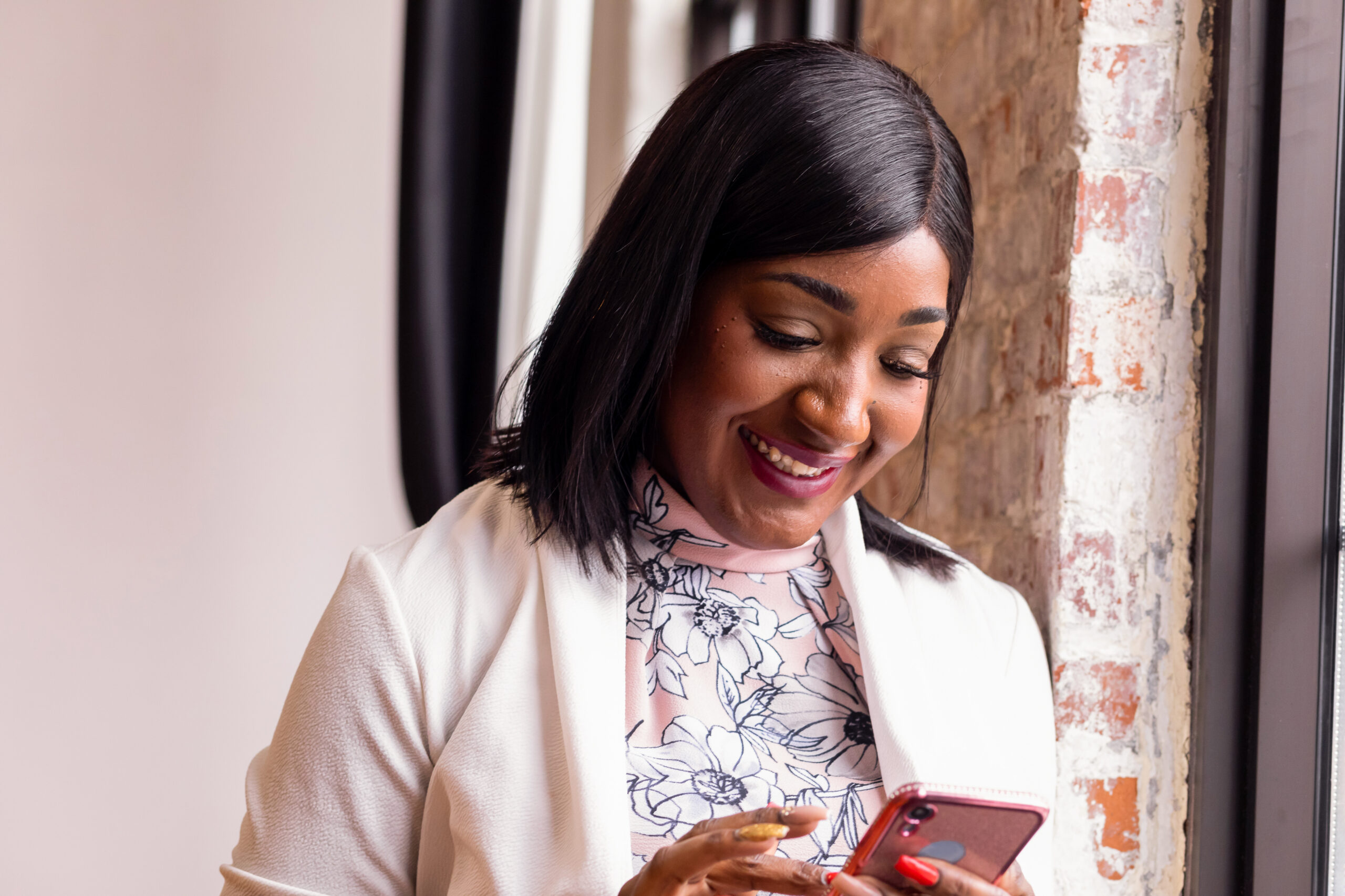 Mulher sorridente, de cabelo preto liso na altura dos ombros, usando um casaco claro sobre uma blusa de gola alta com estampa floral. Ela está próxima a uma janela, com luz natural iluminando o rosto, olhando para um celular que segura com as duas mãos. Ao fundo, há uma parede com tijolos aparentes, criando um ambiente aconchegante e moderno.