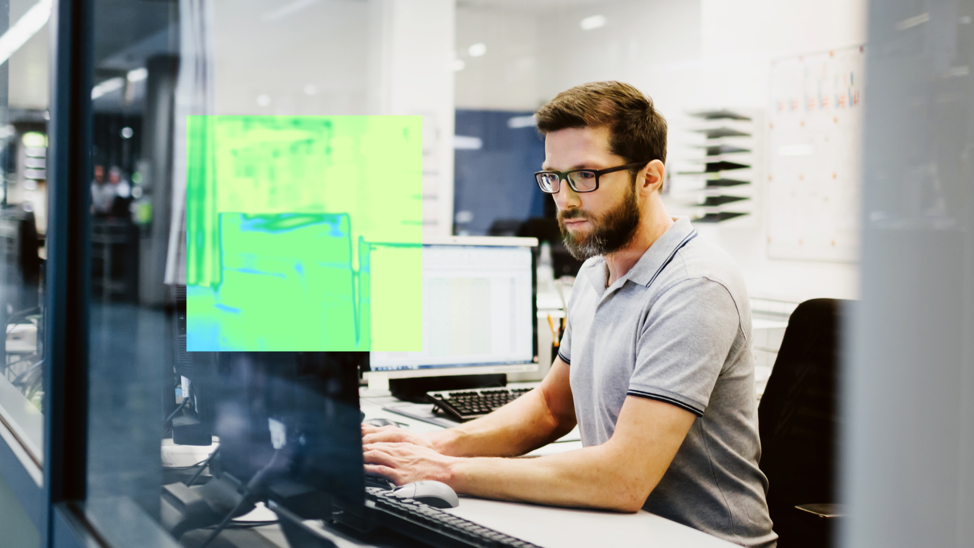 Homem sentado em uma estação de trabalho em um escritório moderno, usando óculos e camisa polo cinza, enquanto digita em um teclado e observa atentamente a tela do computador. À esquerda, há um monitor em primeiro plano com uma sobreposição gráfica em tons de verde e azul, sugerindo uma visualização tecnológica ou interface digital. O ambiente ao fundo é claro, com divisórias de vidro, equipamentos de escritório e uma atmosfera corporativa.